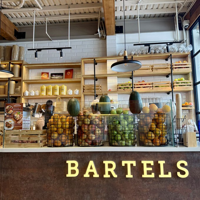 Photo of a juice bar counter labeled 'BARTELS' displaying wire baskets filled with fresh fruits like melons, apples, and oranges, with wooden crates of produce on shelves in the background.