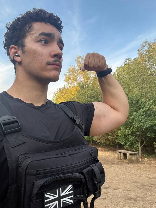 A young man with curly hair and a mustache flexes his bicep while wearing a black tactical chest rig featuring a Union Jack patch, wireless earbuds, and a smartwatch, set against a wooded outdoor backdrop with a stone bench. The image visually reinforces the post's message about tackling an 11k run as a personal challenge tied to PulseChain, with his flexed arm symbolizing the strength and resilience celebrated in the caption.