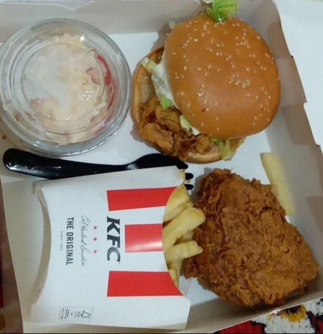 An overhead view of a KFC meal containing a fried chicken sandwich with lettuce, fried chicken pieces, french fries, coleslaw, and a plastic fork in a branded box.
