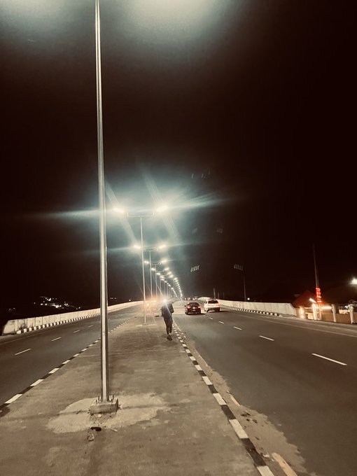 A nighttime photograph of a person walking across a wide empty road or bridge illuminated by tall streetlights, with a few cars visible in the distance.