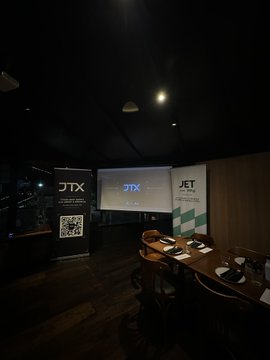 A dimly lit interior of a meeting room or private dining space with multiple screens displaying 'JTX' branding and a QR code, set up with tables and chairs.