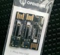 The image features a small black box with three USB memory cards inside, each of which has a gold stripe running down its center. The background appears to be a black surface or cloth, providing contrast for the objects within the frame. The text "OPENDIME" is written in white at the top left corner and "10 years ago OPENDIME was born Bearer Bitcoin Pass it on Keep it safe Today it remains one of the Coolest Bitcoin products".