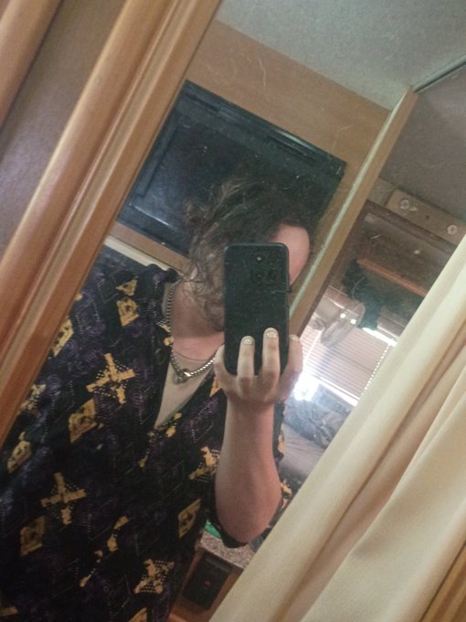 A mirror selfie of a person wearing a dark patterned shirt and silver chain necklace, holding a smartphone to take the photo.