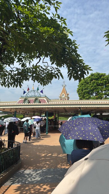 A sunny outdoor scene at what appears to be a theme park entrance with a clock tower and flags, where visitors are walking under a covered walkway, some carrying umbrellas for shade.