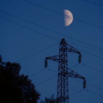 A half moon shines in a deep blue sky above a silhouetted electrical transmission tower and tree tops.