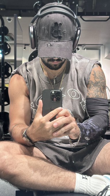 A mirror selfie of a muscular, tattooed man wearing a distressed grey "UC" baseball cap, large headphones, a grey graphic t-shirt, and black shorts, taking a photo with a black smartphone in a gym setting.