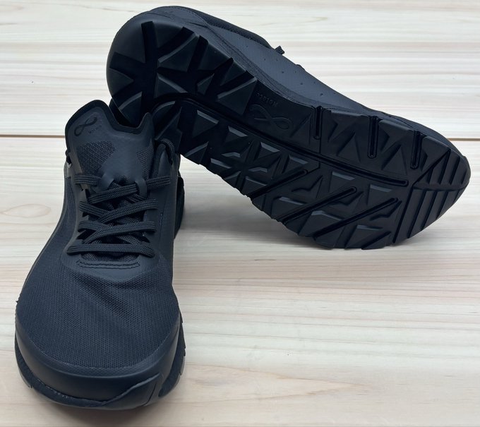 The image shows a pair of black sneakers placed side by side, with their soles facing each other. The sneakers are positioned on a wooden surface, possibly a table or countertop. The caption below the image provides information about the sneakers, stating that they are "StepN" branded shoes and are available in both men's and women's sizes.
