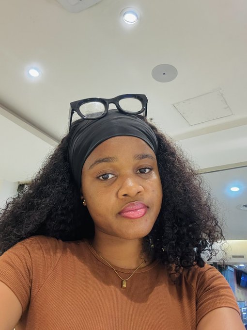 A selfie of a young woman wearing a brown shirt, black headband, and glasses perched on her head, with a neutral expression, complementing the cheerful "GM Have an amazing day" post.