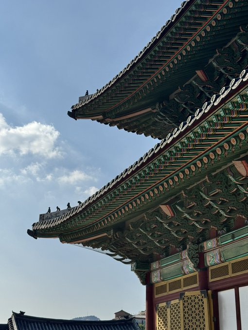 A low-angle photograph of traditional East Asian architecture featuring an ornate, multi-tiered roof with intricate green, red, and gold painted details under a blue sky with scattered clouds.
