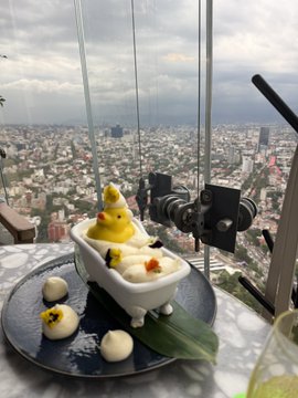 An artistic plated dish featuring yellow chick-shaped dumplings sitting in a tiny white bathtub, accompanied by quail eggs and drizzled sauce, presented on a high-rise balcony overlooking a sprawling cityscape.