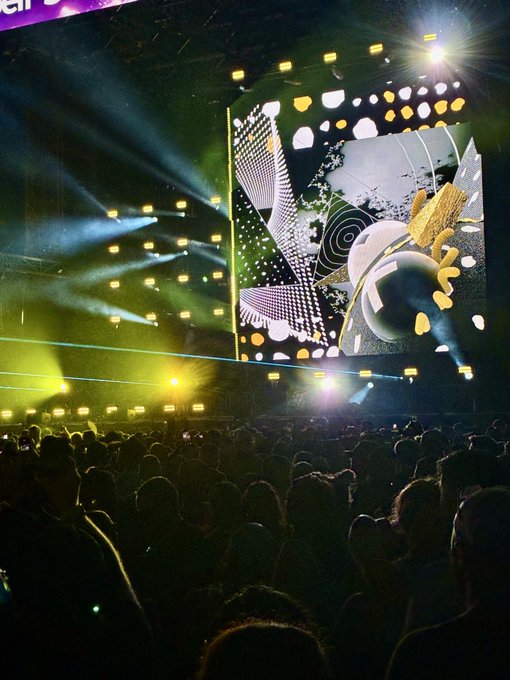 A large crowd gathers at an electronic music concert, illuminated by vibrant yellow and white stage lights with abstract visuals displayed on a massive screen overhead. The dramatic lighting and immersive atmosphere align perfectly with the "Gm Deadmau5" post, capturing the energy of the iconic producer's signature live shows featuring elaborate production design.