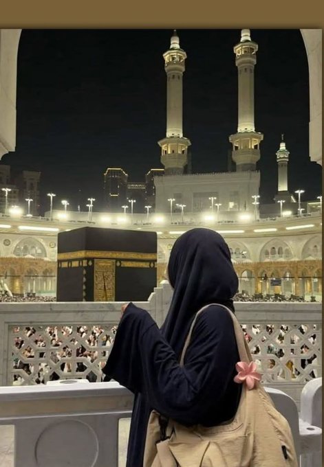 A woman wearing a black niqab and carrying a beige bag stands overlooking the Kaaba at the Masjid al-Haram in Mecca, with the illuminated minarets visible against the night sky.