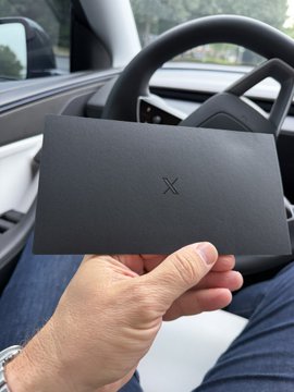 A hand holds up a sleek black metal card featuring a minimalist "X" logo, inside a Tesla with the steering wheel visible in the background. The card matches the post about the @XMoney Card arriving, showing off its premium metallic finish.