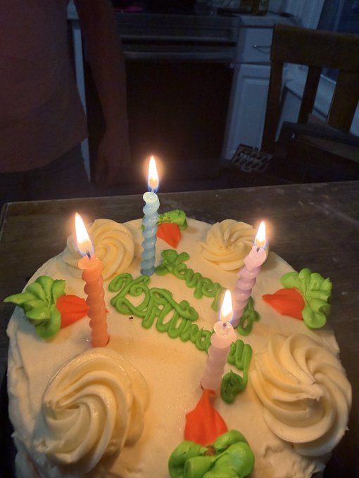 A birthday cake with lit candles and 'Happy Birthday' written in green icing sits on a kitchen counter, with a person standing in the background.