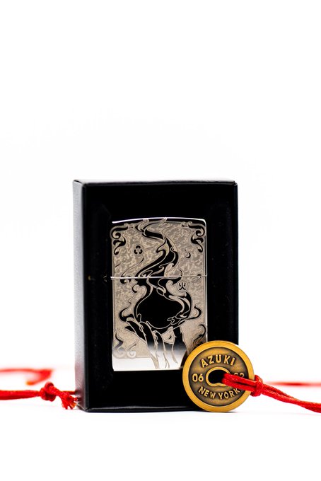 An ornate silver Zippo lighter engraved with a dragon and phoenix design sits in a black presentation box, accompanied by a gold-colored tag reading 'AZUKI 06 NEW YORK' attached with a red string.