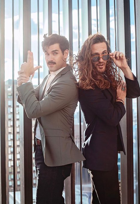 Two men pose back-to-back in suits against a window with city views; the left man has a mustache and gestures with his hand, while the right man has long hair, sunglasses, and a beard.