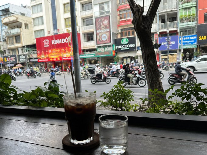 A glass of iced coffee and a glass of water sit on a cafe table overlooking a busy Vietnamese street with motorbikes and storefronts like 'KINH ĐÔ' and 'PHÚC LONG'.