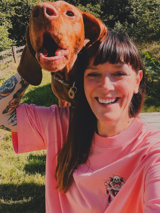A smiling woman in a pink sweatshirt taking a selfie outdoors while holding up a brown dog behind her head, with green trees and grass in the background.