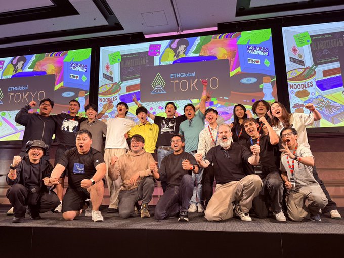 A large group of excited attendees poses for a group photo at the 'ETHGlobal TOKYO' event, holding signs with logos for ETH, Noodles, and EthXY.