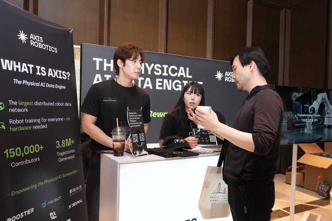 Two booth representatives, a man and a woman in black shirts, speak with an attendee at the Axis Robotics booth, which features banners describing the company as 'The Physical AI Data Engine' with '150,000+ Contributors' and '3.8M+ Trajectories Generated.'