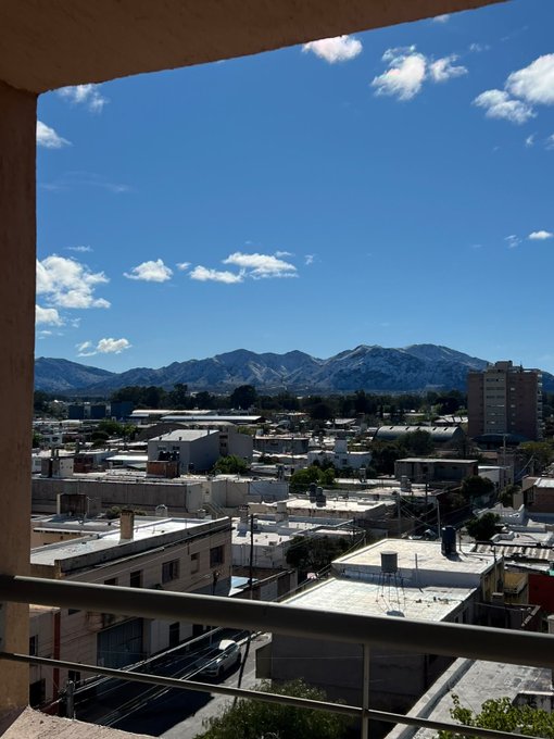 A daytime view from a balcony overlooking a town with low-rise buildings, streets, and a mountain range in the distance under a blue sky with scattered clouds.