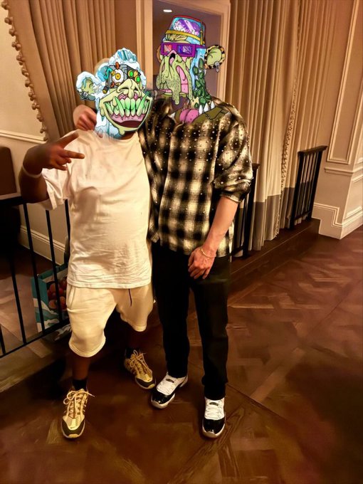 Two people posing together indoors, both with cartoon-style monster face stickers covering their faces, one wearing a white t-shirt and shorts, the other in a black and white plaid shirt.