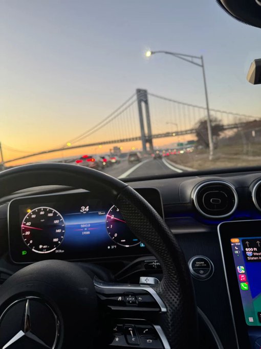 A view from the driver's seat of a Mercedes-Benz cruising toward the Verrazzano-Narrows Bridge at dusk, with the dashboard showing "34" mph and navigation indicating "500 ft" to the Verrazzano Bridge—matching the "GM" (Good Morning) request with a scenic morning commute shot.