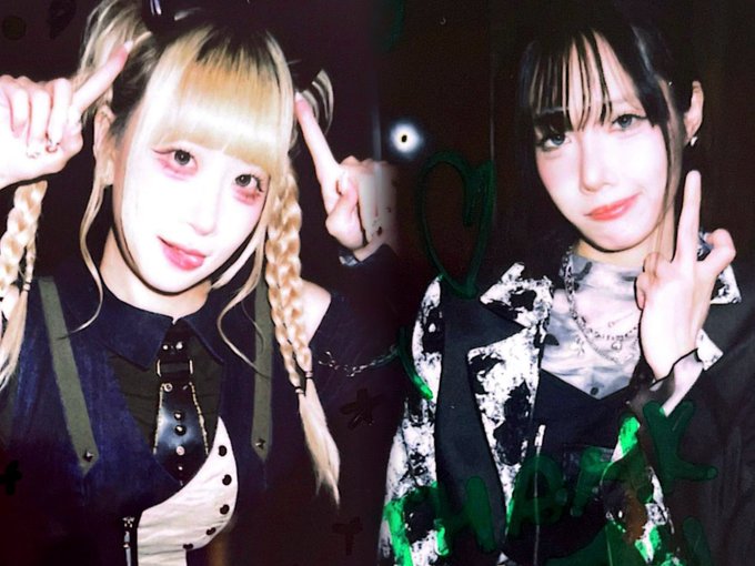 Two young women pose in a dimly lit setting, one with blonde braids and the other with dark hair, both making peace signs and wearing dark, edgy outfits.