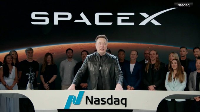 Elon Musk stands at a Nasdaq podium during a SpaceX event, with the 'SPACEX' logo visible in the background and 'Nasdaq' displayed on the podium.