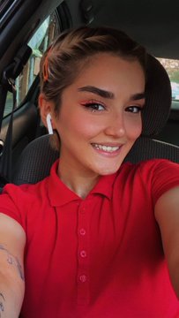 A woman with dark hair pulled back takes a selfie in a car, wearing a red polo shirt, smiling with bright makeup and wireless earbuds.