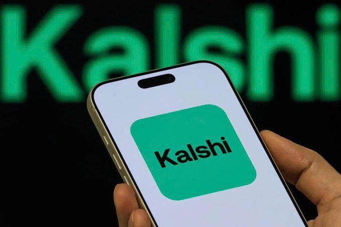A hand holding a smartphone displaying the green 'Kalshi' app icon, with the brand name blurred in the background.