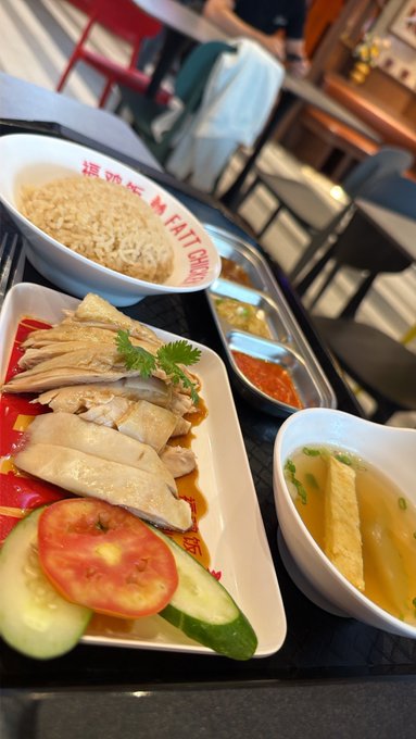 A plate of Hainanese chicken with rice is served at Fatt Choy Eating House, featuring sliced poached chicken garnished with cilantro, accompanied by tomato and cucumber slices, soup with bamboo pith, and chili sauce.
