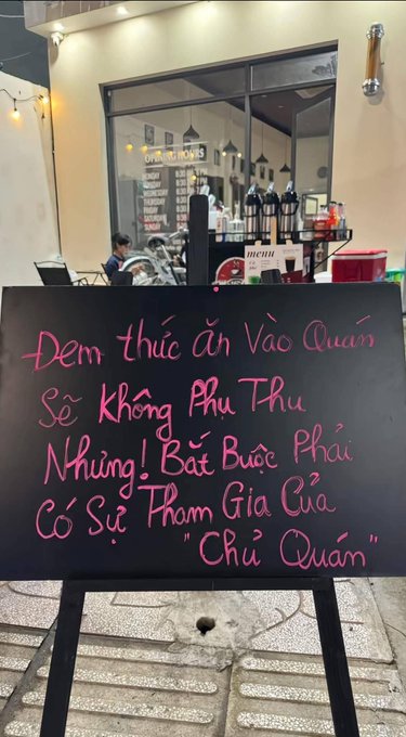 A black chalkboard sign with pink Vietnamese handwriting displayed outside a restaurant reads: 'Đem thức ăn vào quán sẽ không phụ thu, nhưng! Bắt buộc phải có sự tham gia của "Chủ Quán"' (bringing outside food incurs no surcharge, but the owner's participation is required).