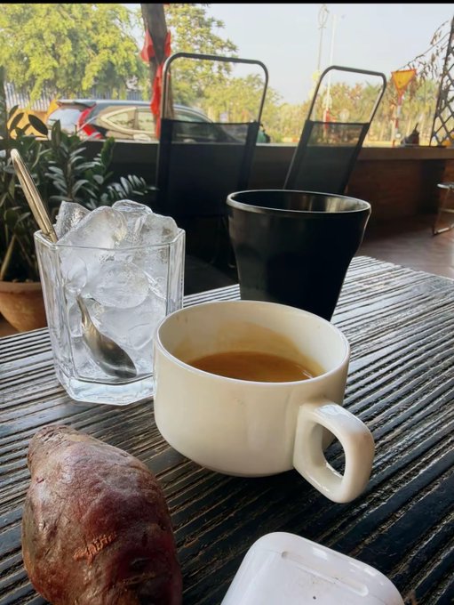A close-up of a café table setting with a white ceramic coffee cup containing espresso, a black coffee cup, a glass filled with ice and a spoon, and a sweet potato, with outdoor seating and trees visible in the background.