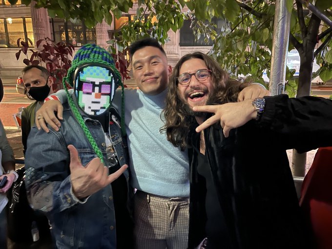 Three people pose outdoors at night, smiling and making hand gestures, with one wearing a green knit hat and an LED pixel-art face mask, while another wears a light blue turtleneck and the third has long hair and glasses.
