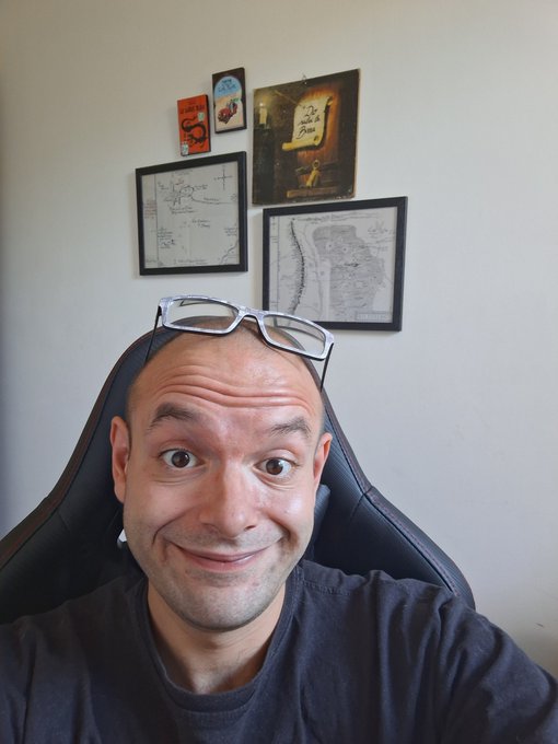 A bald man with a big smile and a wide-eyed, excited expression sits in a gaming chair, with white-framed glasses perched on his head. Behind him, the wall features framed artwork including a "Seth Godin" book cover, a "Do the Work" print, and two framed maps. The cheerful selfie matches the upbeat, energetic tone of the post announcing a packed Wednesday schedule filled with gaming streams, videos, and crypto activities.