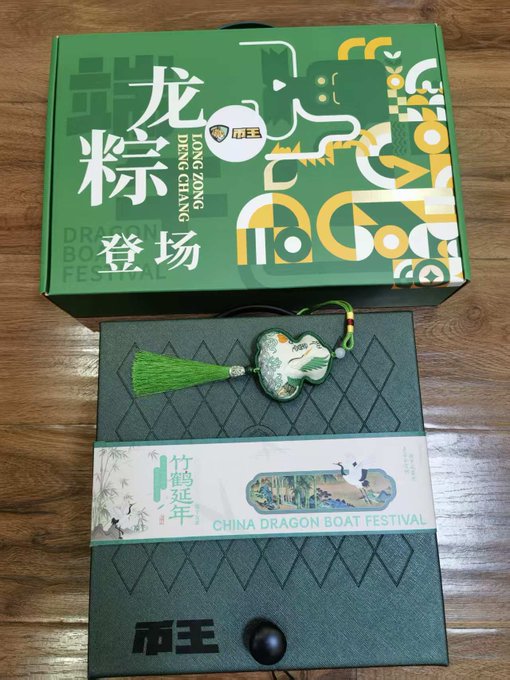 The image features two boxes, one green with Chinese characters and a dragon boat festival logo, and another box that appears to be a gift box or a package. The boxes are placed next to each other on a wooden surface. The text "收到币王 @BiKing_CN 的端午节礼物。茶具一套咸鸭蛋都塞进去了，仪式感到位。 最近戒酒，正好一边喝茶一边看世界杯。". The boxes seem to be related to a Chinese festival or event, possibly featuring dragon boat motifs.
The image also contains text in both English and Chinese that reads "收到币王 @BiKing_CN 的端午节礼物。茶具一套咸鸭蛋都塞进去了，仪式感到位。最近戒酒，正好一边喝茶一边看世界杯".