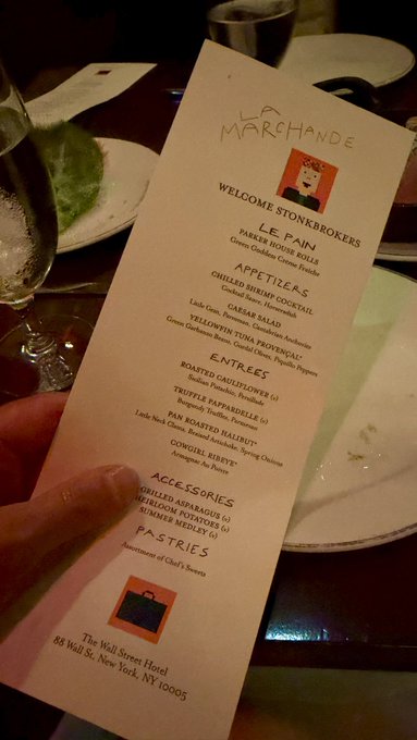 A hand holds a menu for 'LA MARCHANDE' restaurant at The Wall Street Hotel, displaying sections for Appetizers, Entrees, and Pastries.