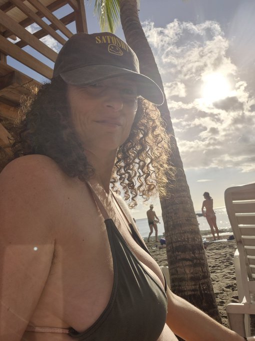 A woman with curly hair wearing a 'SATURDAY' cap and dark bikini top sits at a beach with palm trees and bright sun behind her.
