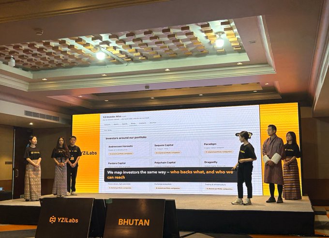 YZi Labs founders demo on stage at a Bhutan event, with a presentation slide listing portfolio investors (Sequoia Capital, Paradigm, Polychain Capital, Dragonfly) and the tagline "We map investors the same way – who backs what, and who we can reach."
