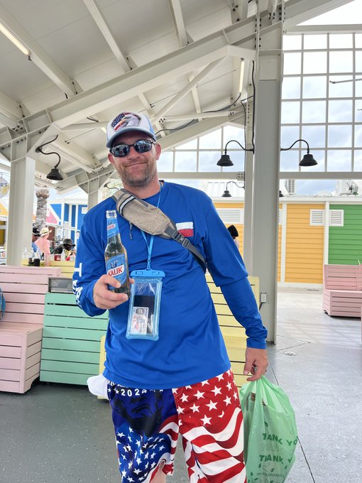 A man wearing a blue long-sleeve shirt, American flag-patterned shorts, and a 'Trump' cap holds a bottle of Michelob Ultra, with a water-resistant phone pouch around his neck, standing under a covered pavilion.