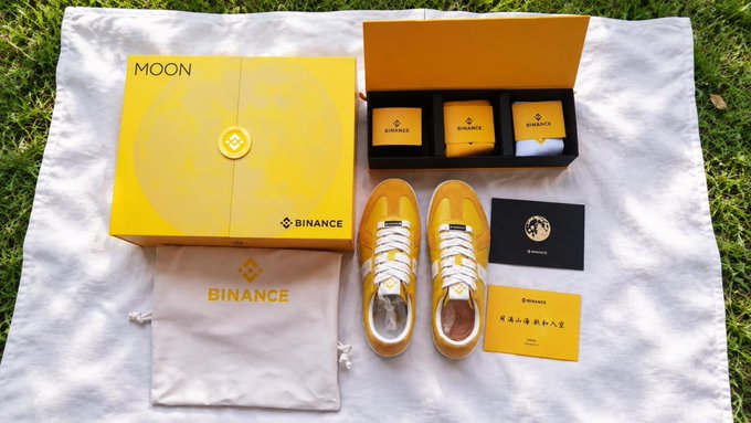 A Binance branded promotional set laid out on grass, featuring a yellow "MOON" box, yellow sneakers, and smaller boxes with Binance logos.