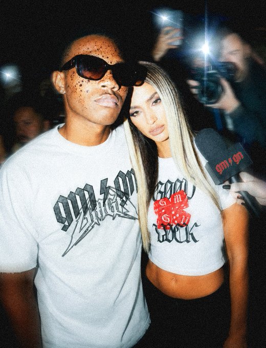 A man wearing dark sunglasses and a 'Vicious' graphic t-shirt poses closely with a blonde woman in a matching 'Vicious' crop top, with someone holding a microphone nearby and another person taking a photo with a flash in the background.