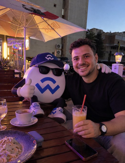 A man smiling and posing at an outdoor cafe table next to a white mascot ball wearing a blue cap and sunglasses with a blue logo, giving a thumbs-up.