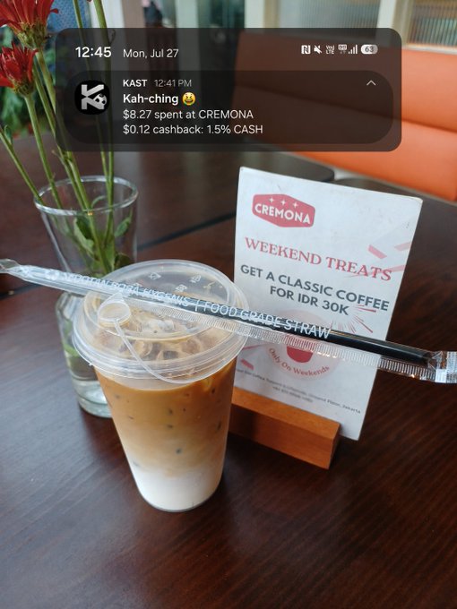 An iced coffee in a clear cup rests on a table beside a 'CREMONA WEEKEND TREATS' sign offering a classic coffee for IDR 30K, with a KAST app notification showing a $0.27 cashback from Kah-ching.