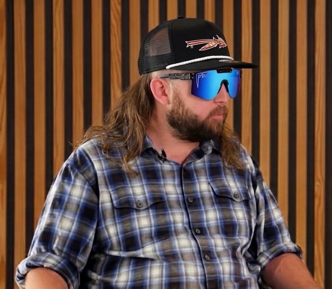 A man with long hair, a beard, and blue reflective sunglasses wearing a trucker hat and blue plaid shirt, sitting in front of a wooden slat wall.