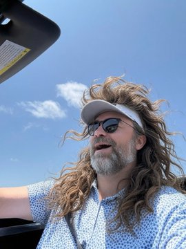 A man with long curly hair, a beard, sunglasses, and a white visor smiles while sitting in a vehicle under a blue sky.