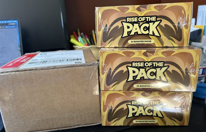 Three identical gold and brown boxes labeled "RISE OF THE PACK" containing 24 booster packs each are stacked on a desk next to a plain brown cardboard shipping box.