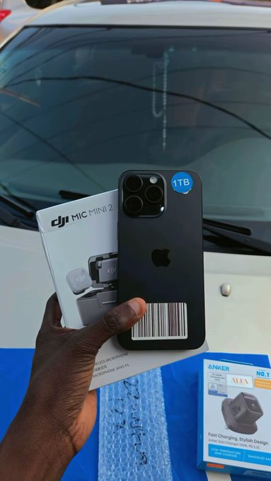 A person holds a DJI Mini 2 drone box and an iPhone 15 Pro Max (1TB) in front of a car, with an Anker charger box visible. The image showcases the tech gadgets purchased by converting cryptocurrency to Naira using the Kazzapp, highlighting a low-fee $1 transaction for over $600 worth of goods.