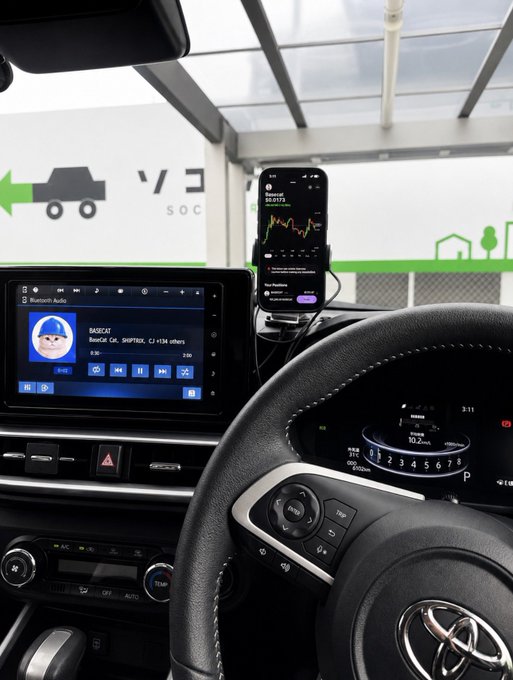 View from the driver's seat of a Toyota vehicle showing the infotainment system playing 'BASSCAT' on Bluetooth Audio, with a smartphone mounted displaying a trading chart for 'Beastly BNB' cryptocurrency.